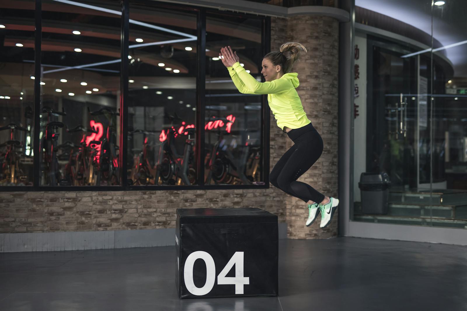 Fit young woman in sports wear executing a box jump indoors, promoting fitness and health in a modern urban gym.